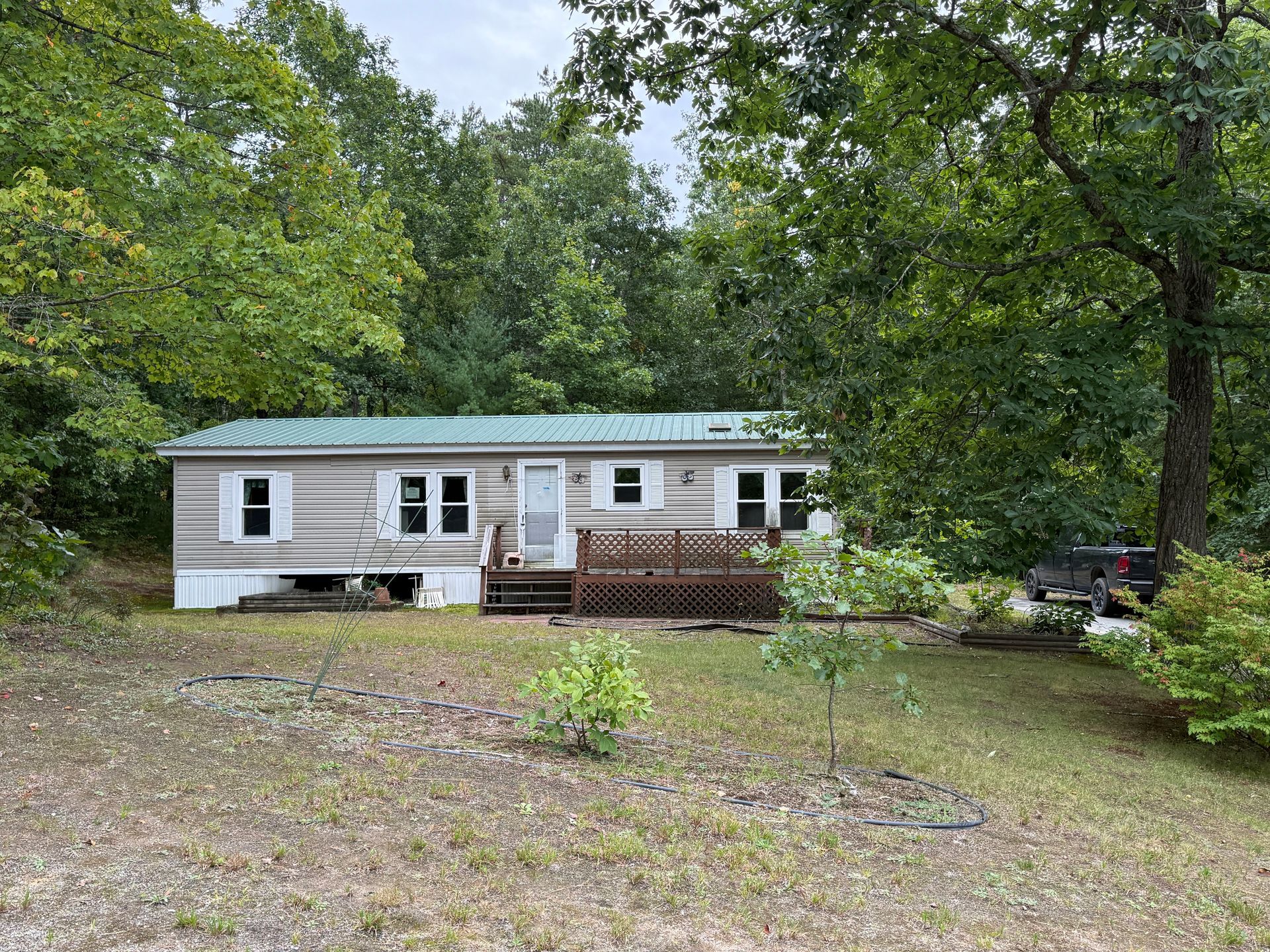 Single-story white house with green roof in a wooded yard, with a small porch and gravel driveway.