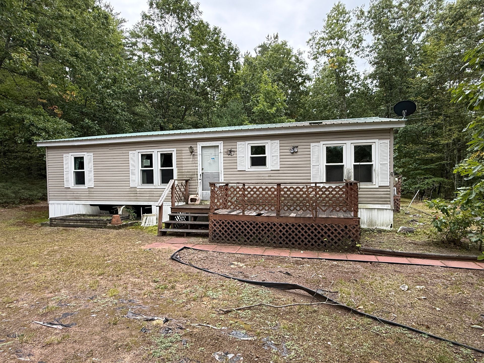 Single-story beige mobile home with a small wooden deck, surrounded by trees and gravel yard.