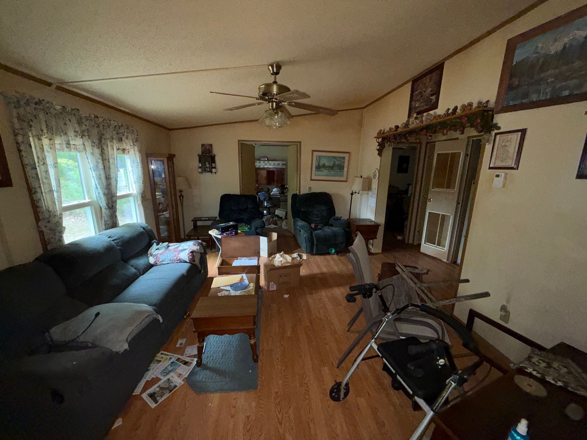 Living room with dark couch, wooden table, recliner chairs, walker, and large windows with curtains.