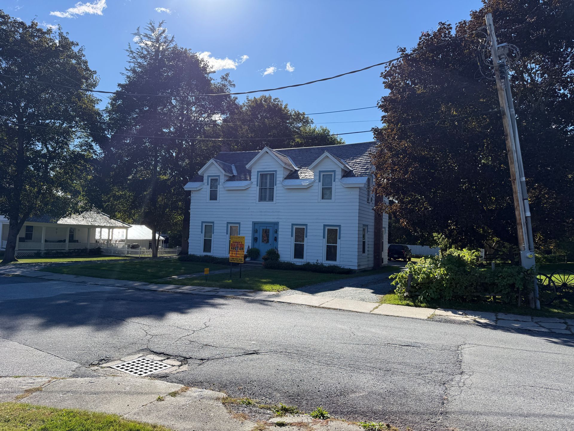 White two-story house with dormer windows on a sunny street, framed by trees and power lines.