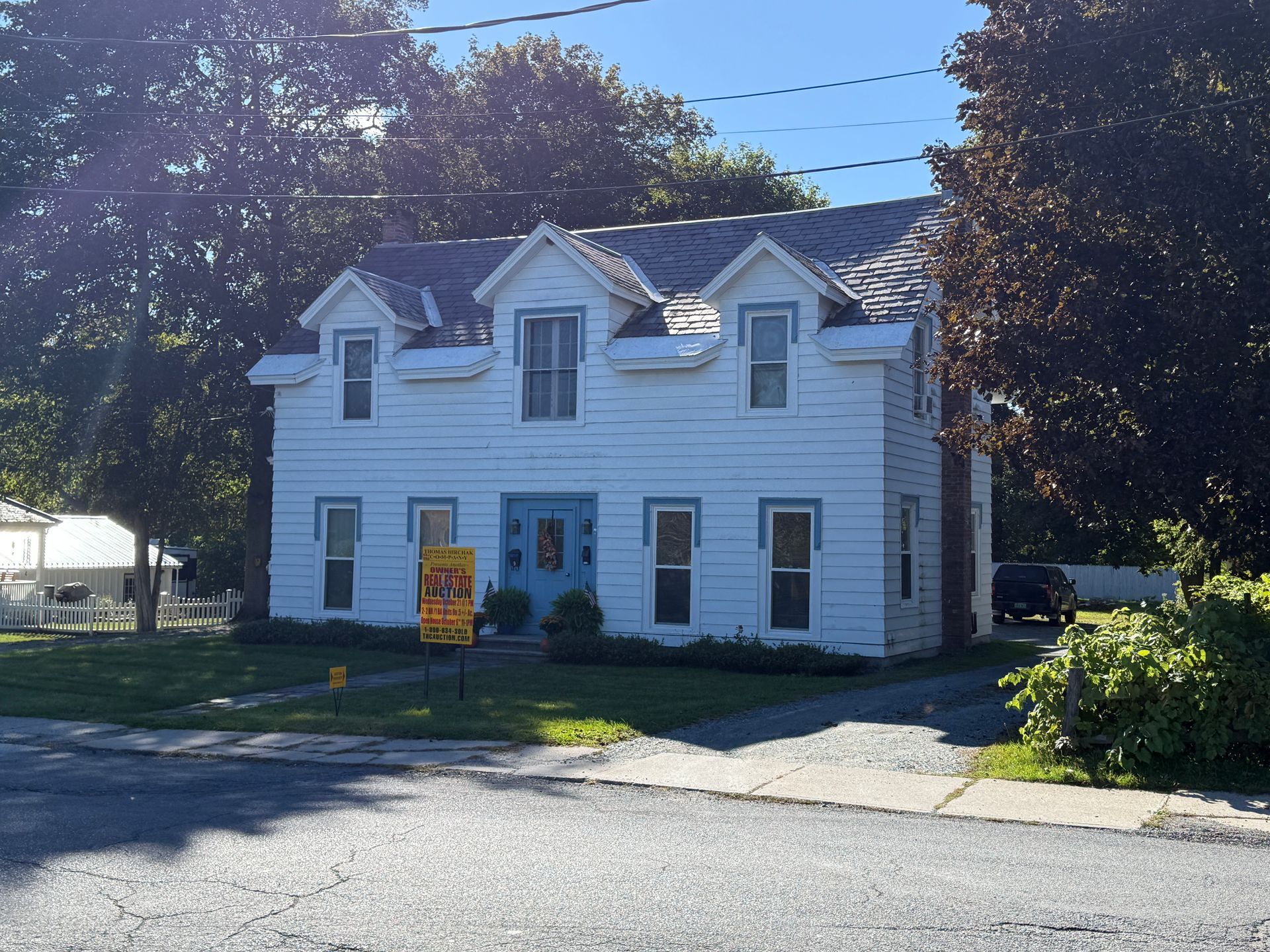 White two-story house with blue trim and a “For Sale” sign in front, shaded by trees