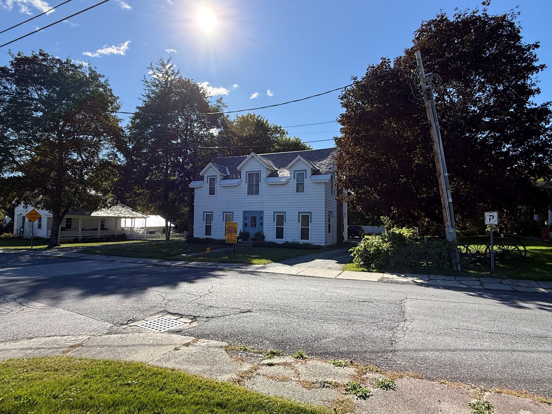 White house with dormer windows on a sunny street, framed by trees and overhead utility lines.