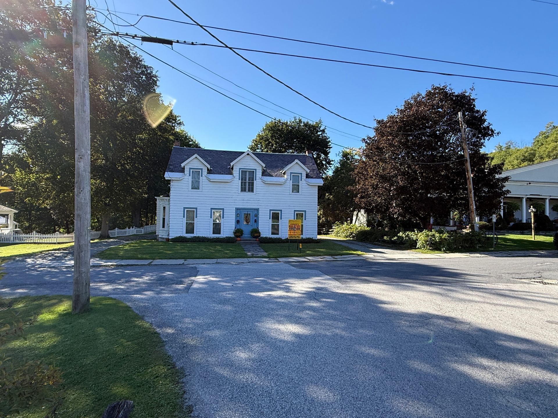 White two-story house with blue shutters on a sunny street, viewed across a gravel lot.