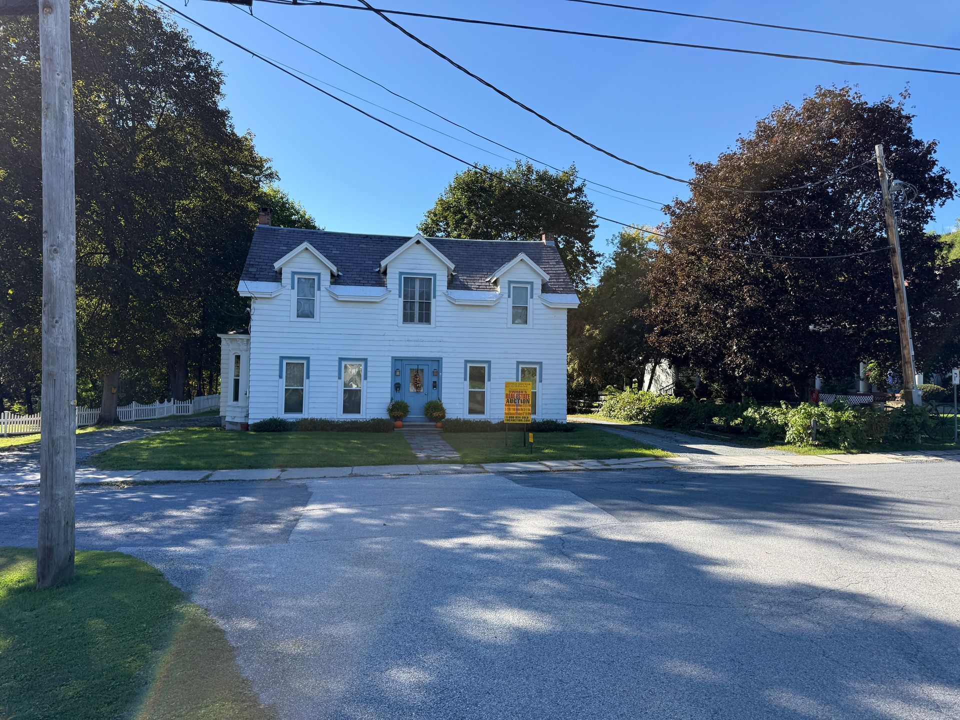 White two-story house with dormer windows and blue shutters behind a gravel driveway and trees