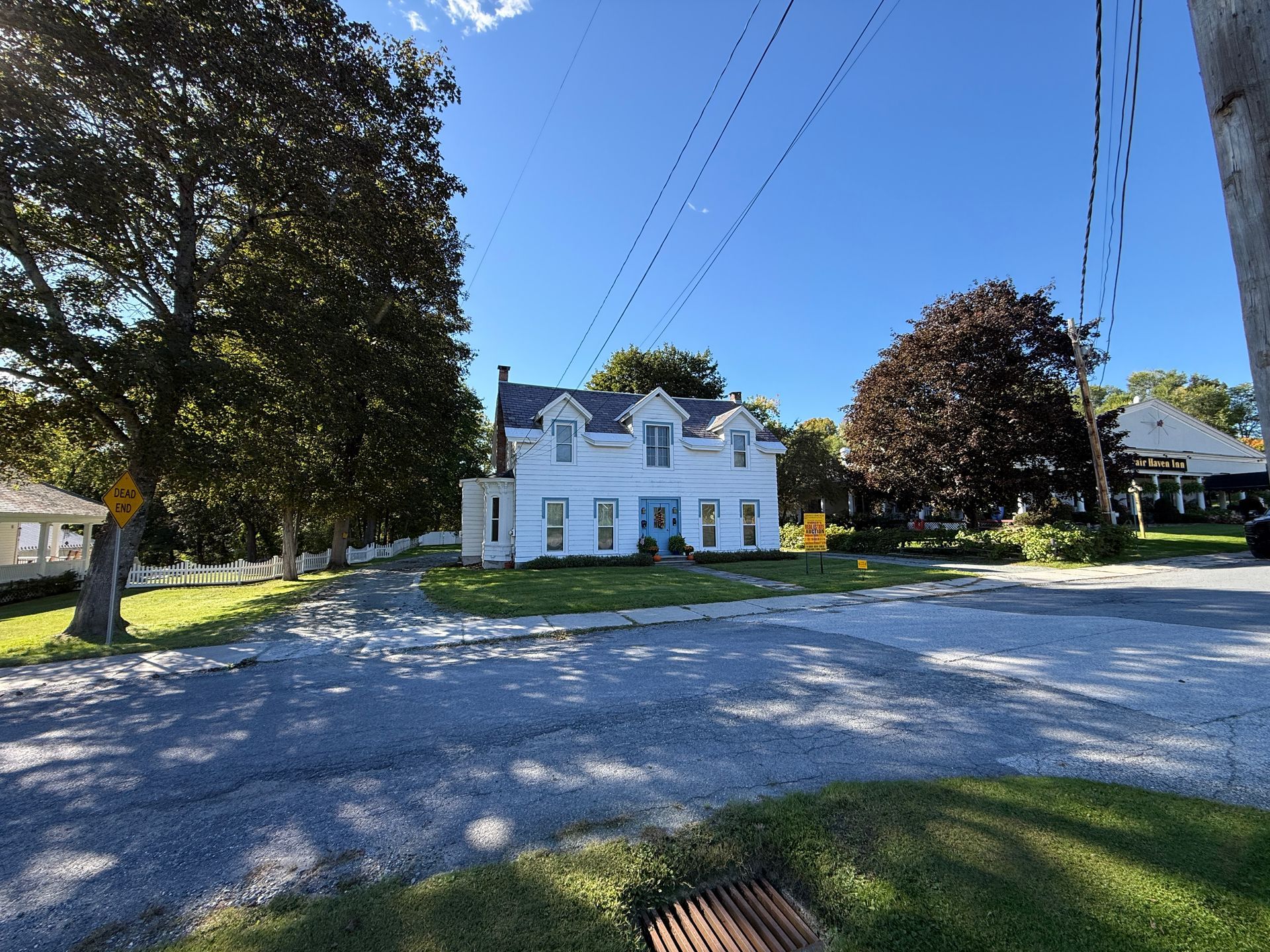 White farmhouse with green shutters beside a gravel lot, framed by trees under a clear blue sky.