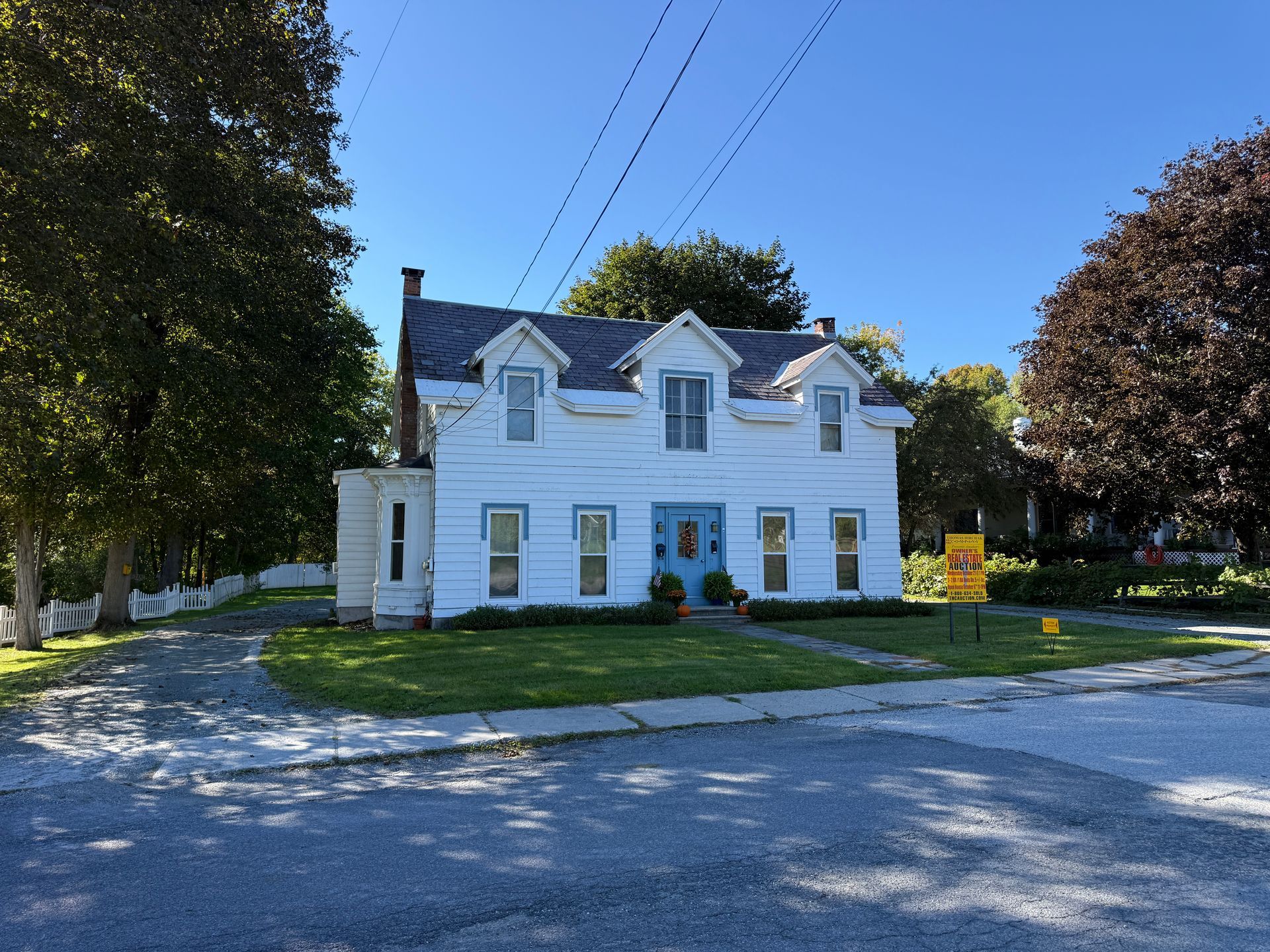 White two-story house with dormers, front lawn, and trees beside a road under a clear blue sky