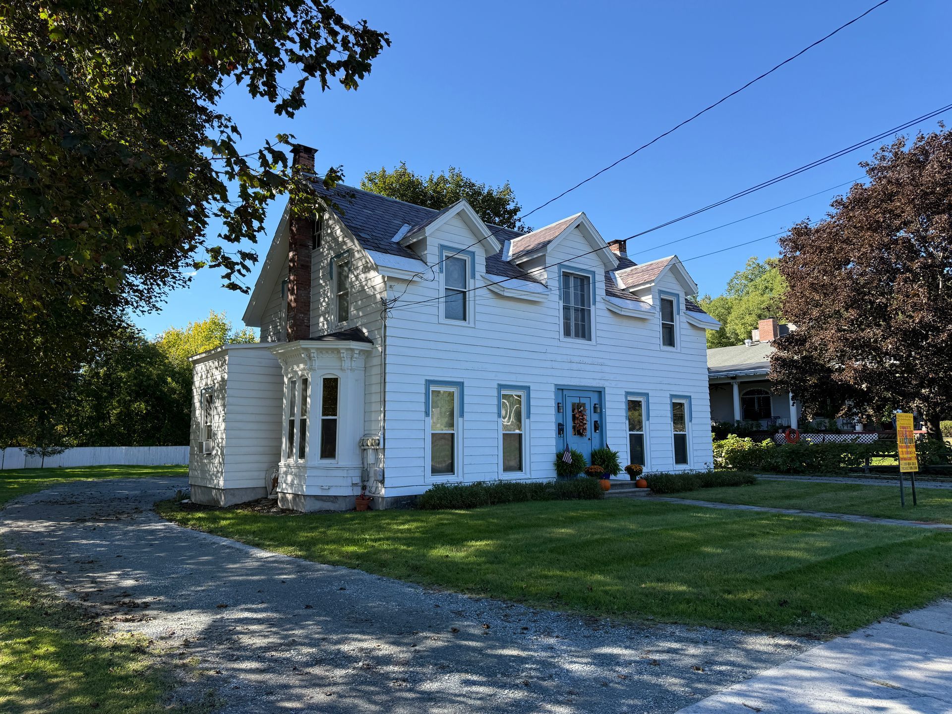 White two-story house with dormer windows and a small porch on a sunny tree-lined street.