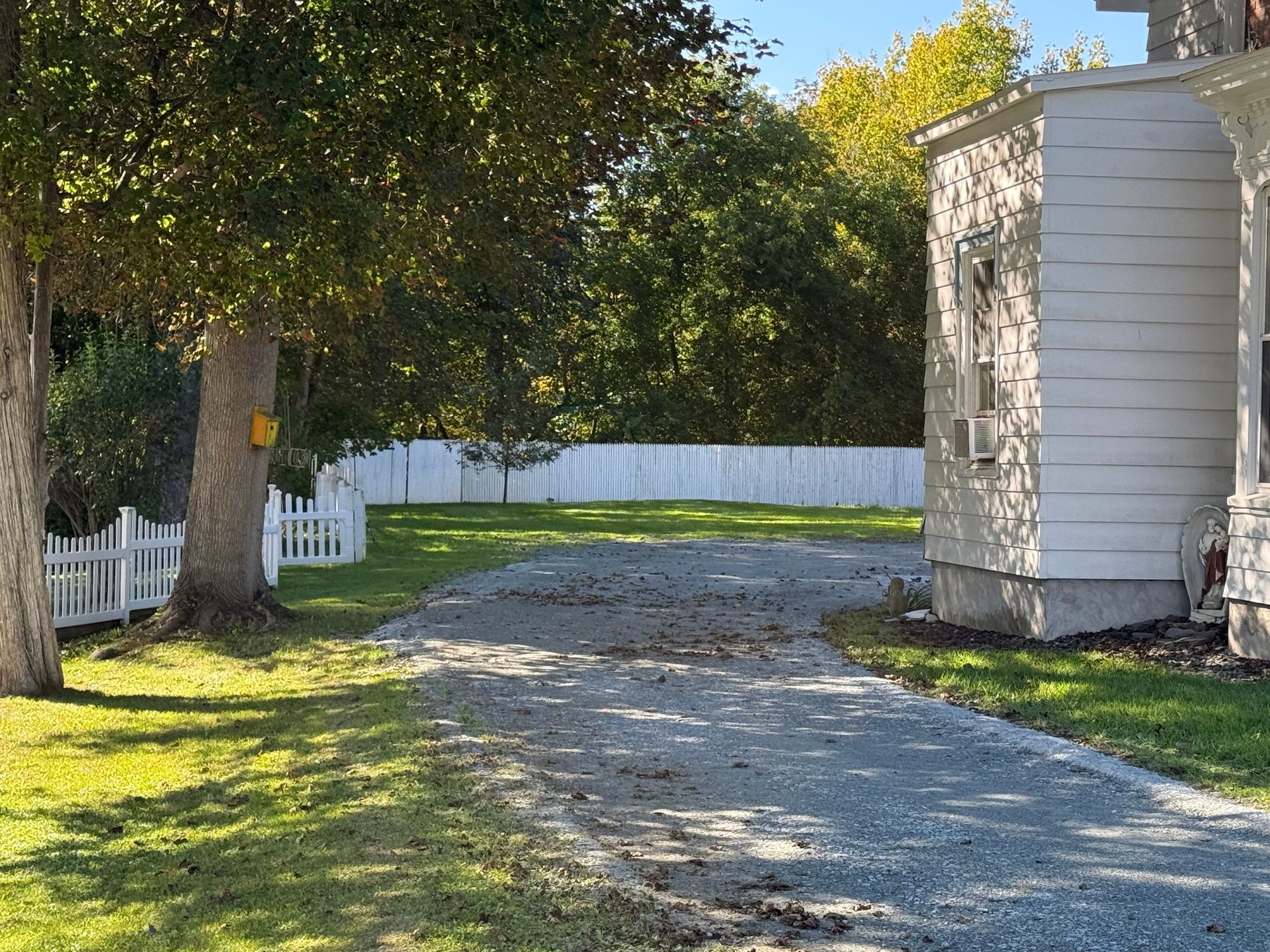 Gravel driveway beside a white house and fence, shaded by trees on a sunny day.