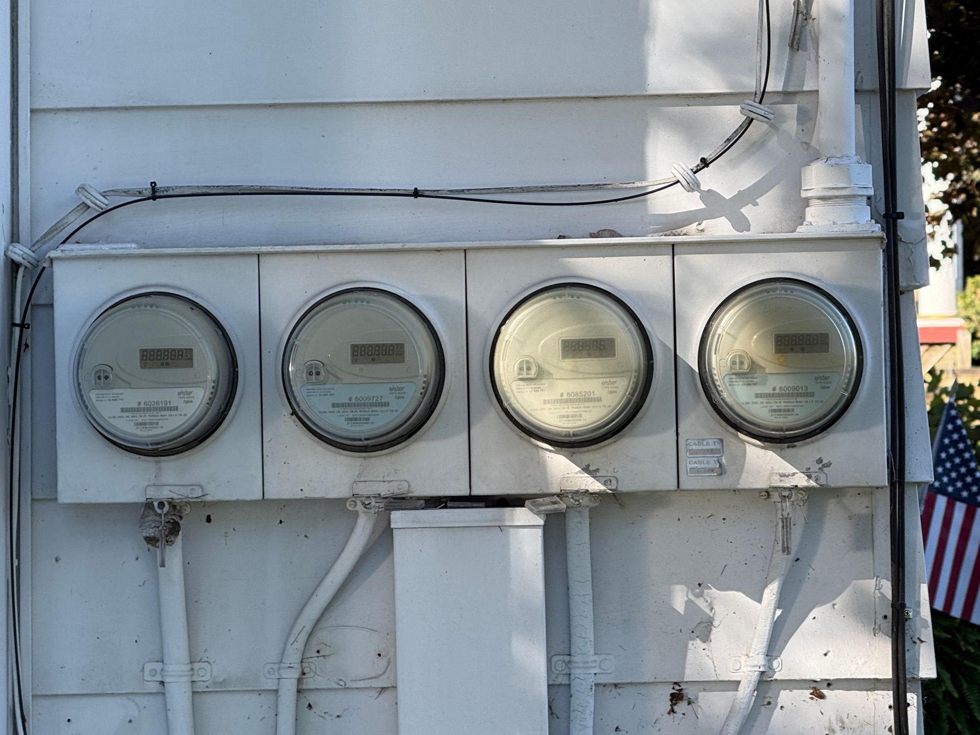 Four round electric utility meters mounted on a white exterior wall under cables