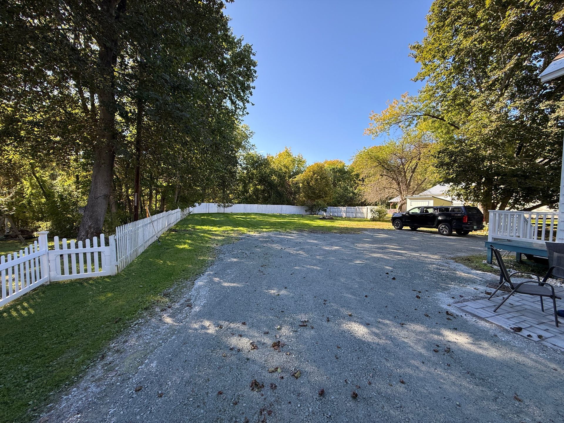 Gravel driveway lined with white fences and trees, with a parked black SUV under a blue sky
