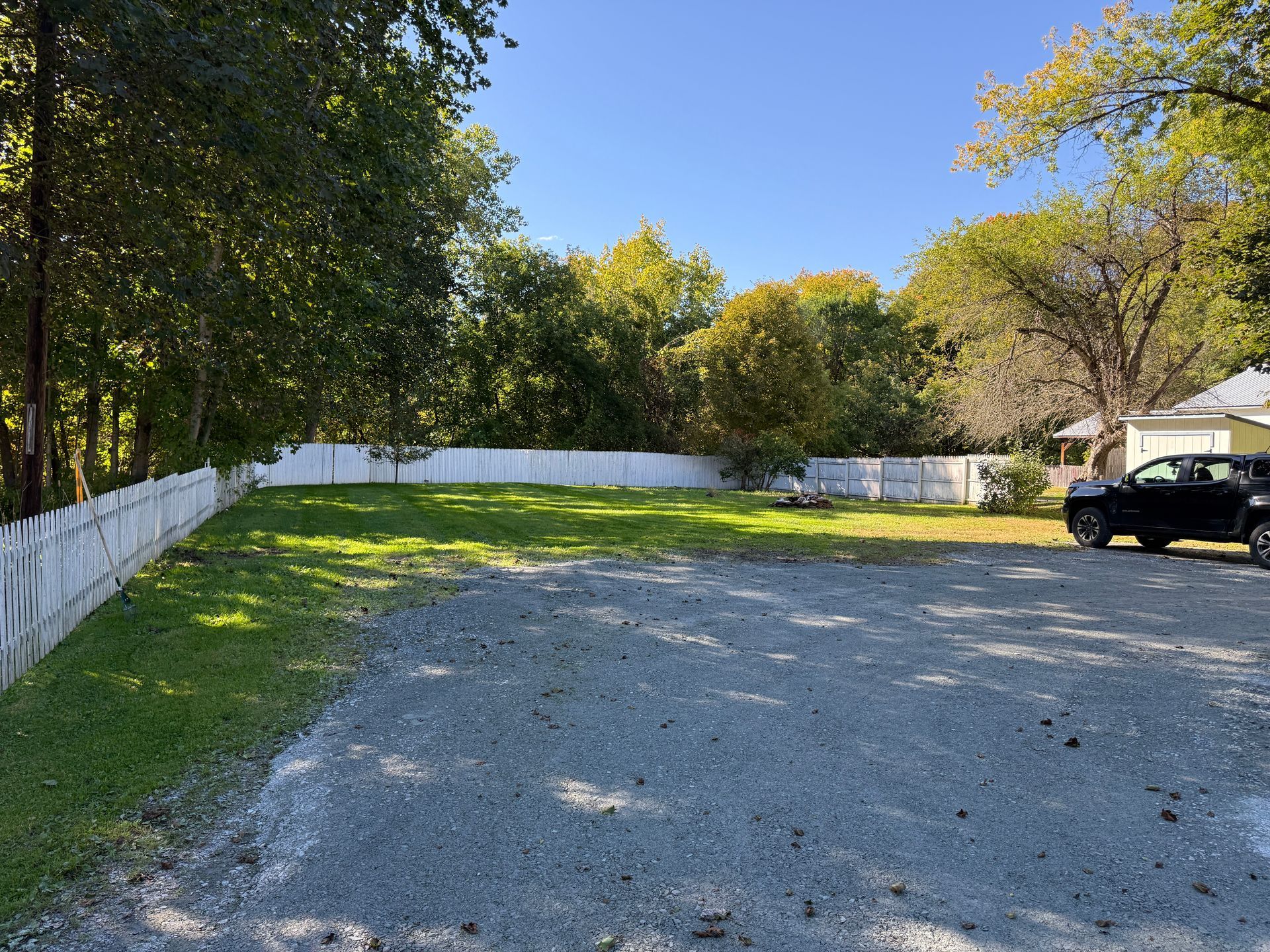Gravel parking lot beside a white fence and trees, with a black pickup truck at the edge