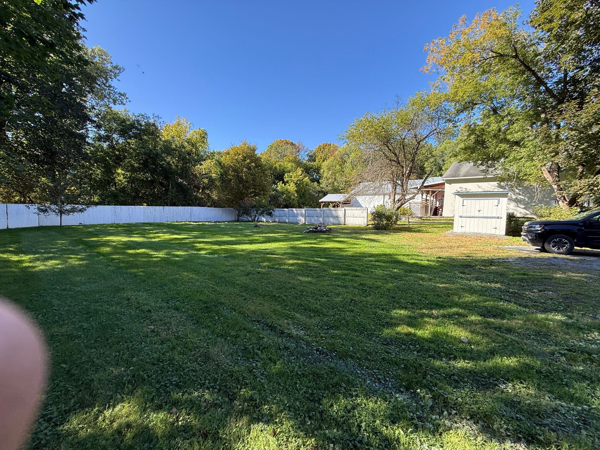 Sunny backyard with green lawn, white fence, trees, and a house with a parked car on the right.