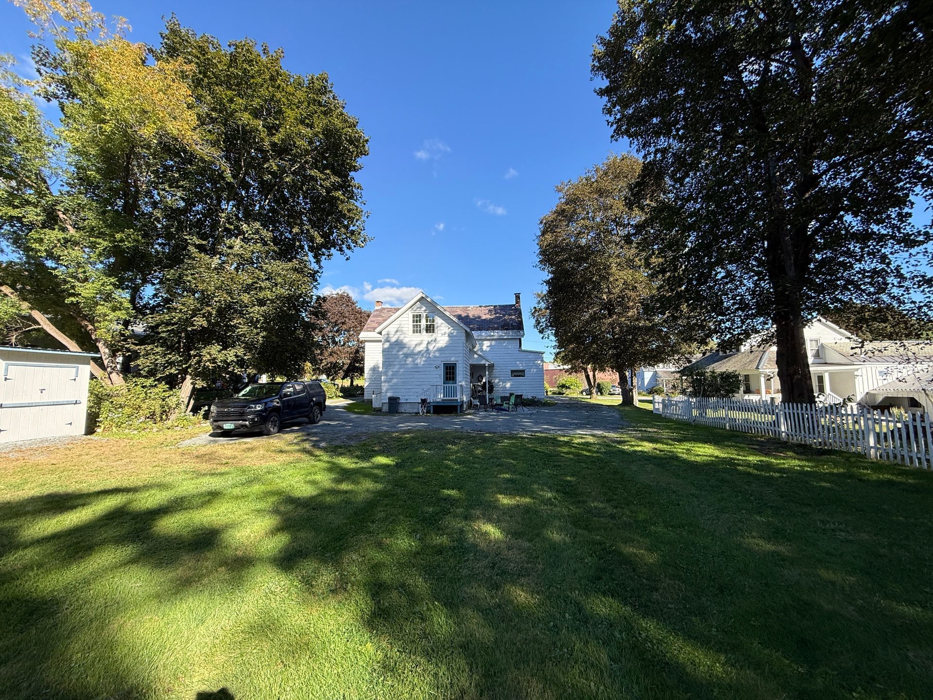 White house with porch in a sunny grassy yard, framed by trees under a clear blue sky.