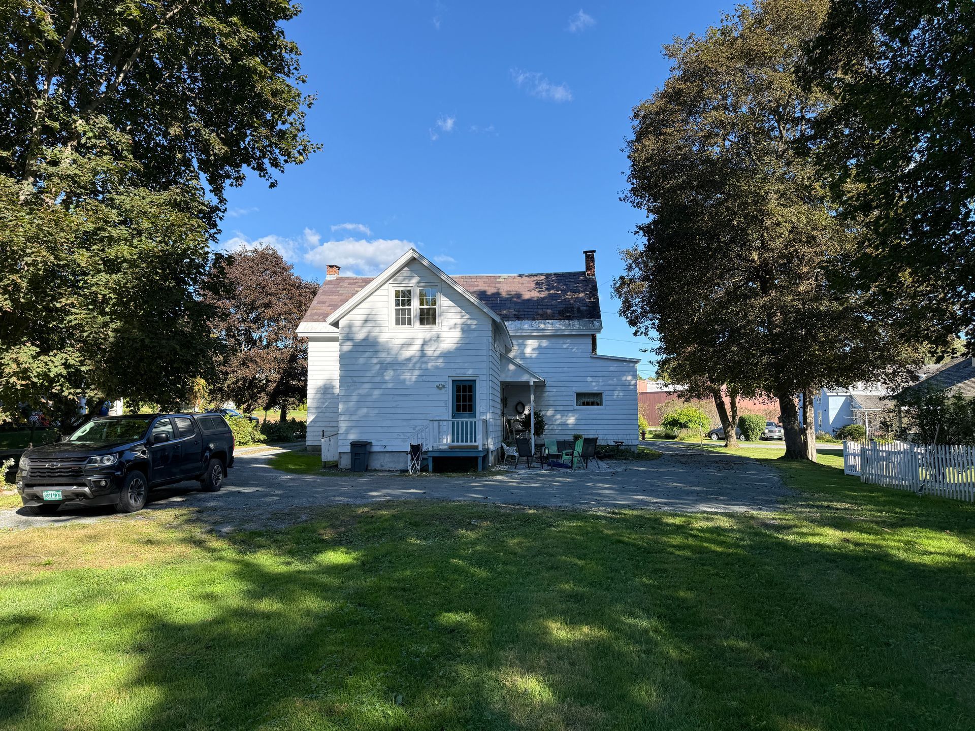 White two-story house with a black car in the driveway, framed by trees and a green lawn under a blue sky