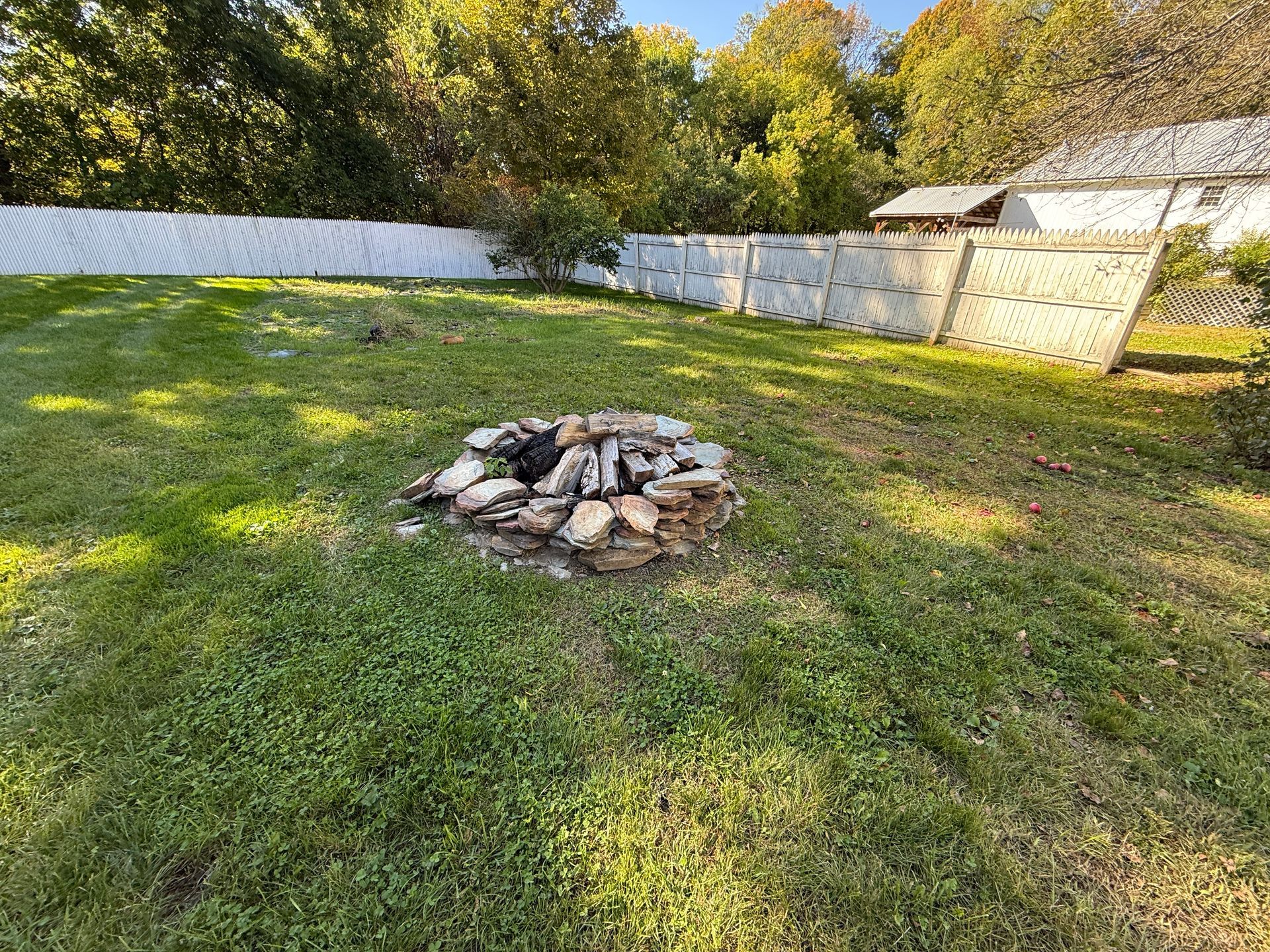 Backyard lawn with a stone fire pit centered near a white fence and trees in sunlight