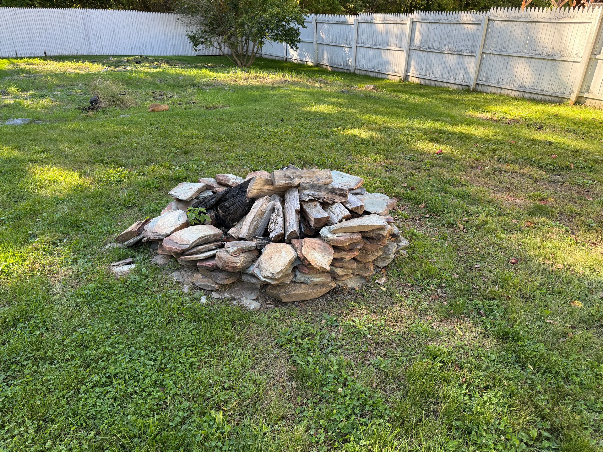 Small stone fire pit in a grassy backyard near a white fence.