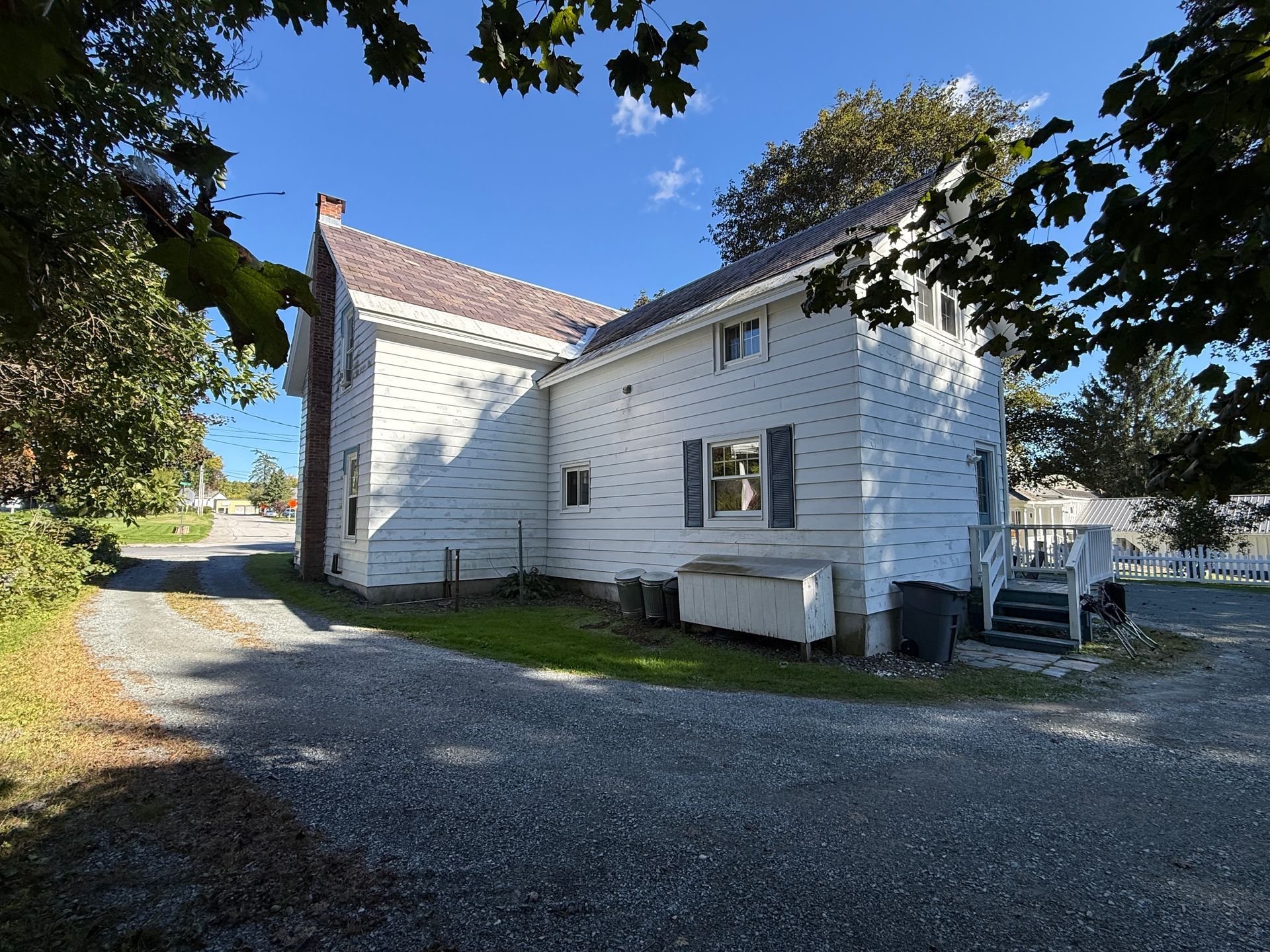 White wooden house with red roof beside a gravel driveway, framed by trees under a blue sky