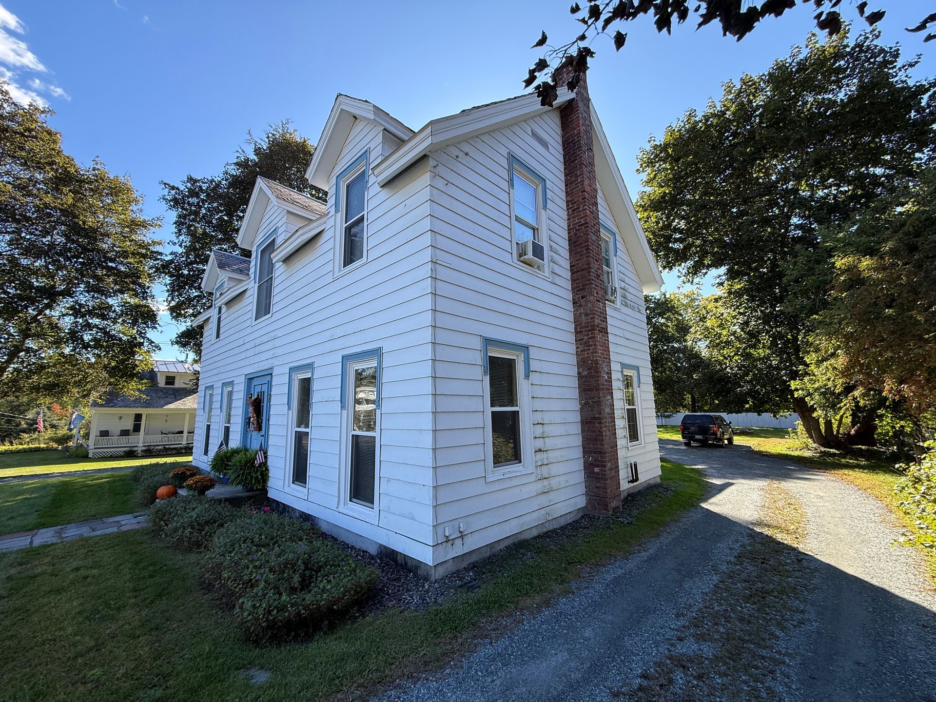 White two-story house with dormer windows and brick chimney, viewed from a grassy driveway under a sunny sky