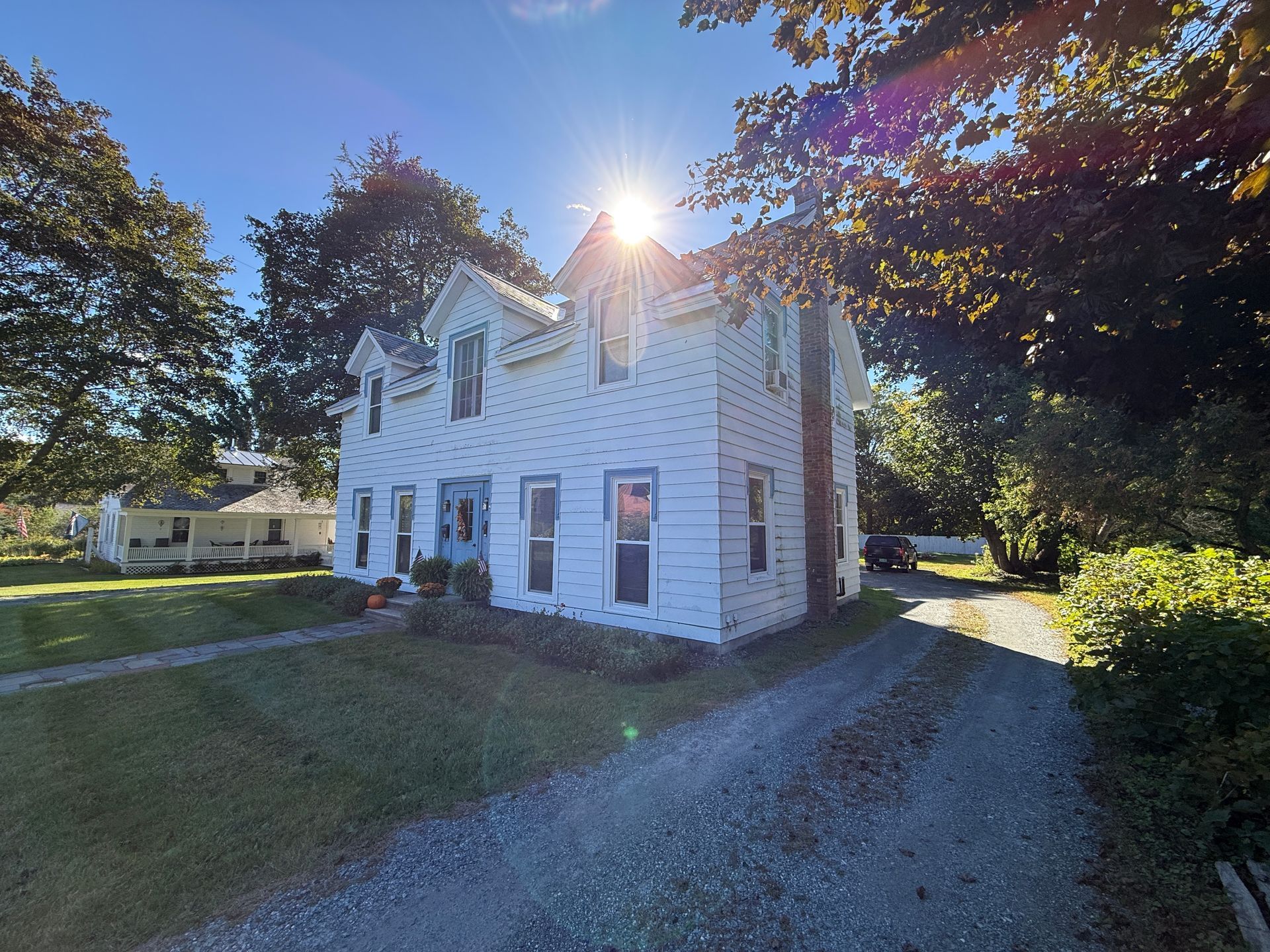 White two-story house with sunlit roof, gravel driveway, and trees in the yard