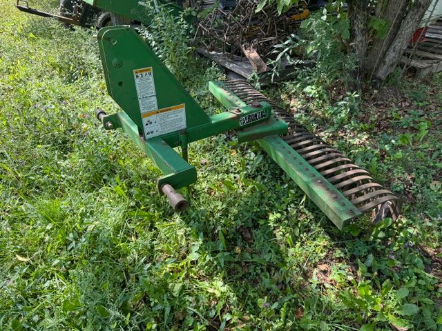 Green farm tiller attachment resting in tall grass beside a wooded area