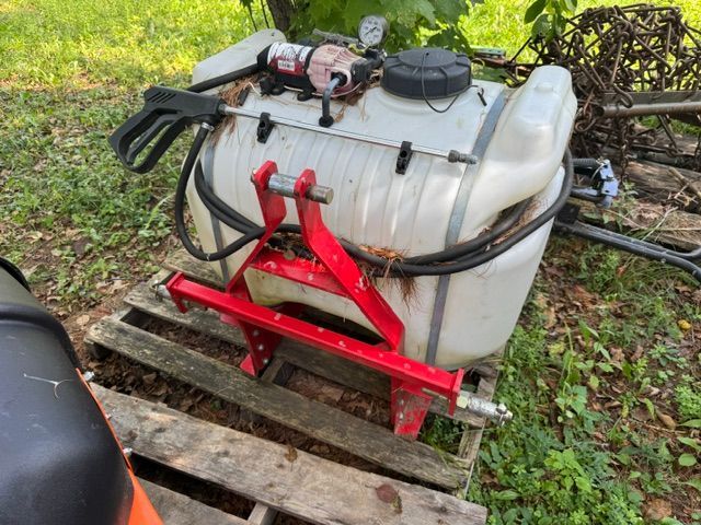 White tank sprayer on a red frame cart, sitting on a wooden pallet outdoors in grass