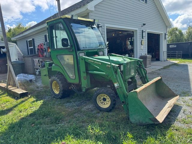 Green utility tractor with front bucket parked beside a garage in a grassy yard.
