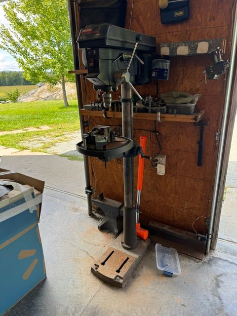 Drill press mounted on a wheeled base in a garage, with outdoor light visible through the open door.