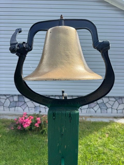 Large bronze bell mounted on a black metal frame in a grassy yard beside a building