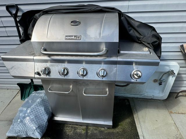 Stainless steel gas grill covered with a black tarp on a patio against a metal wall.