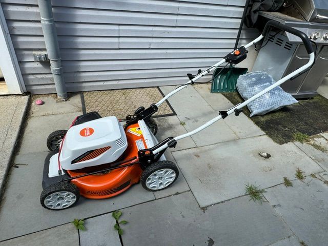 Orange-and-white push lawn mower on a concrete driveway beside a garage door, next to trash bins.