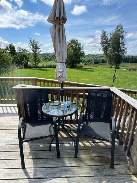 Patio deck with black chairs, glass table, closed umbrella, and a view of a green lawn and trees.
