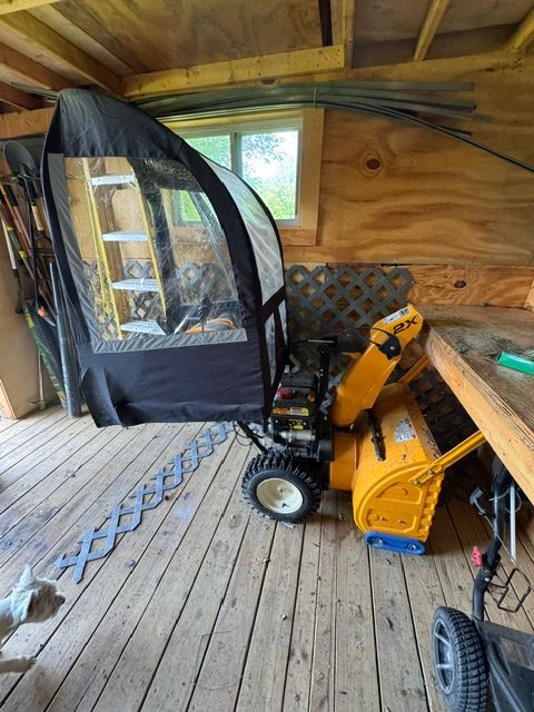 Yellow snowblower in a wooden shed beside a black tent-like enclosure and a window.