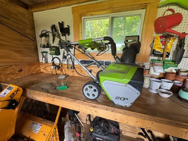 Green and gray snow blower on a wooden workbench in a small shed workshop