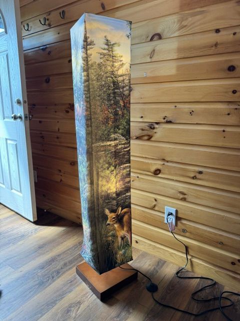 Tall painted floor lamp with forest scene beside a wood-paneled wall and doorway