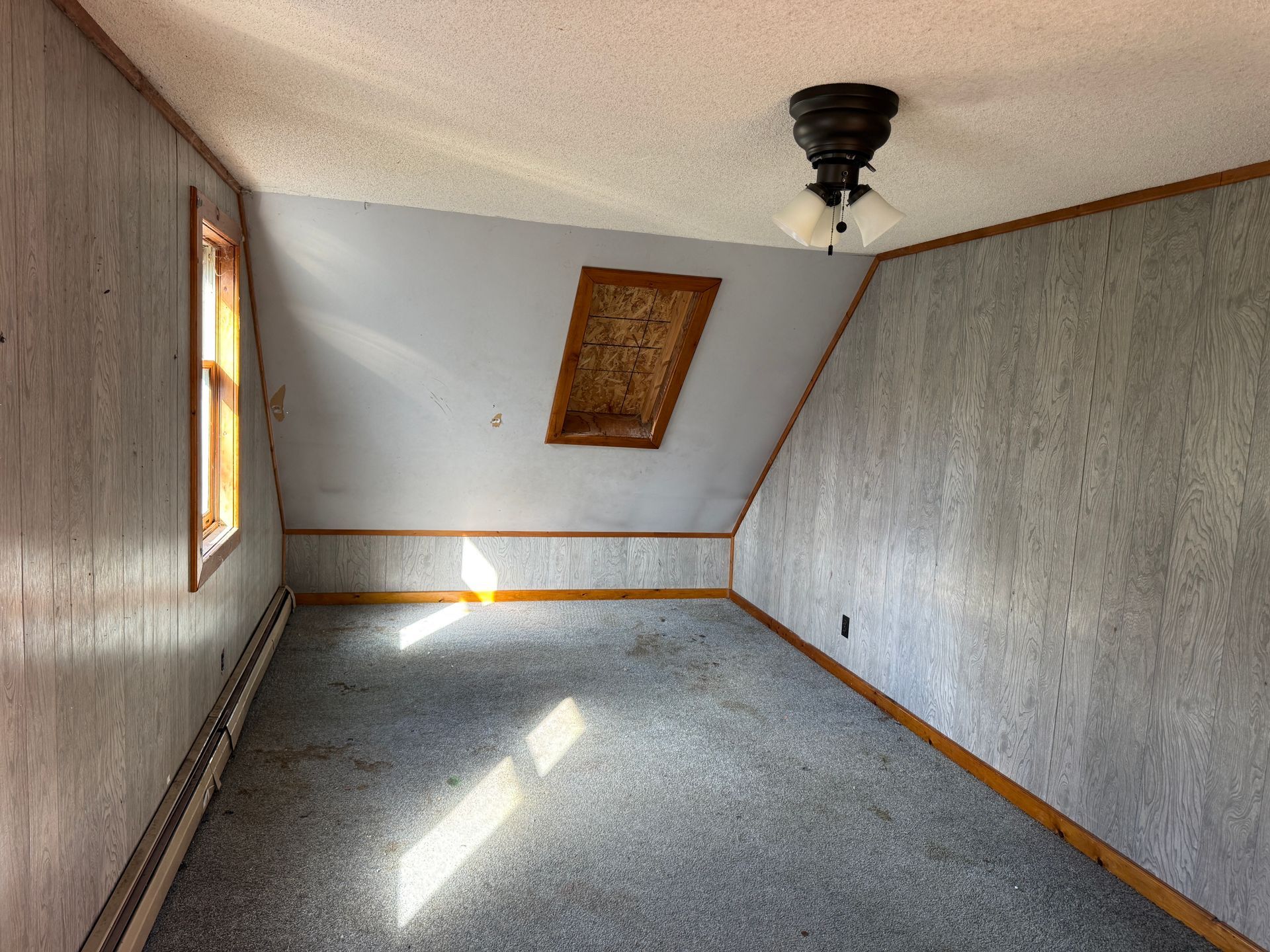 Empty slanted room with gray carpet, gray walls, a small window, and sunlight on the floor.