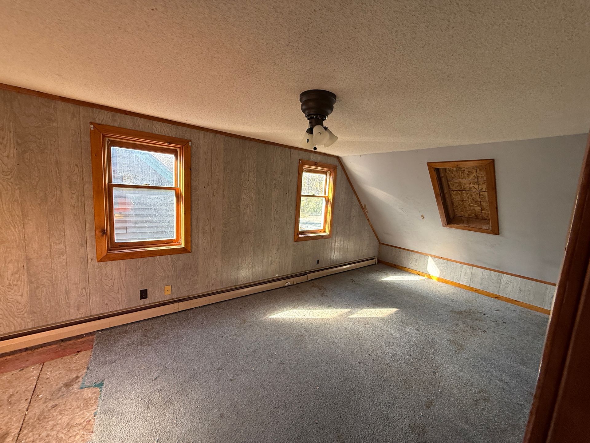 Empty attic room with gray carpet, wood trim windows, slanted ceiling, and hanging light fixture