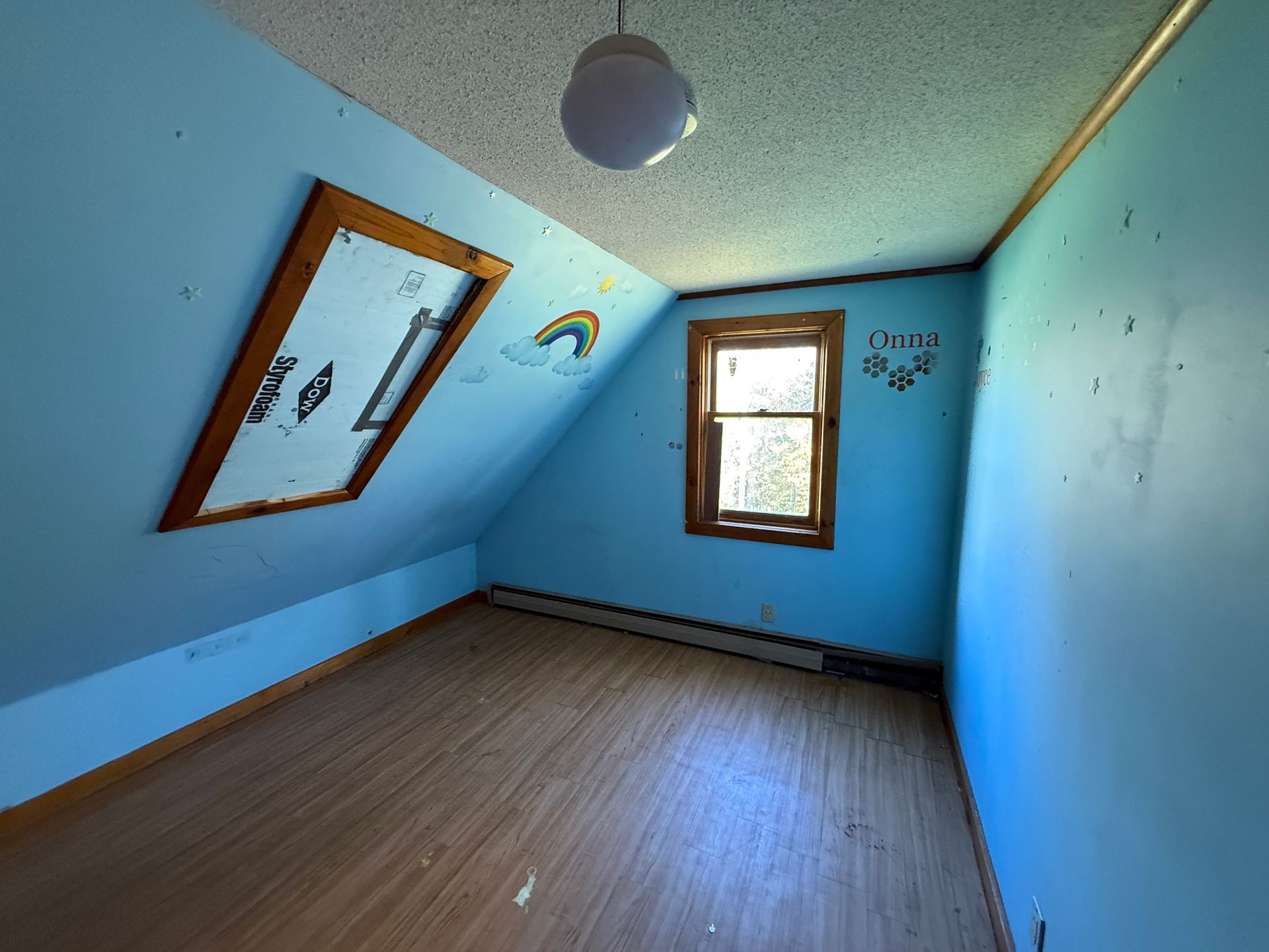 Empty blue bedroom with slanted walls, a window, and a ceiling light; mirror leans against the left wall.