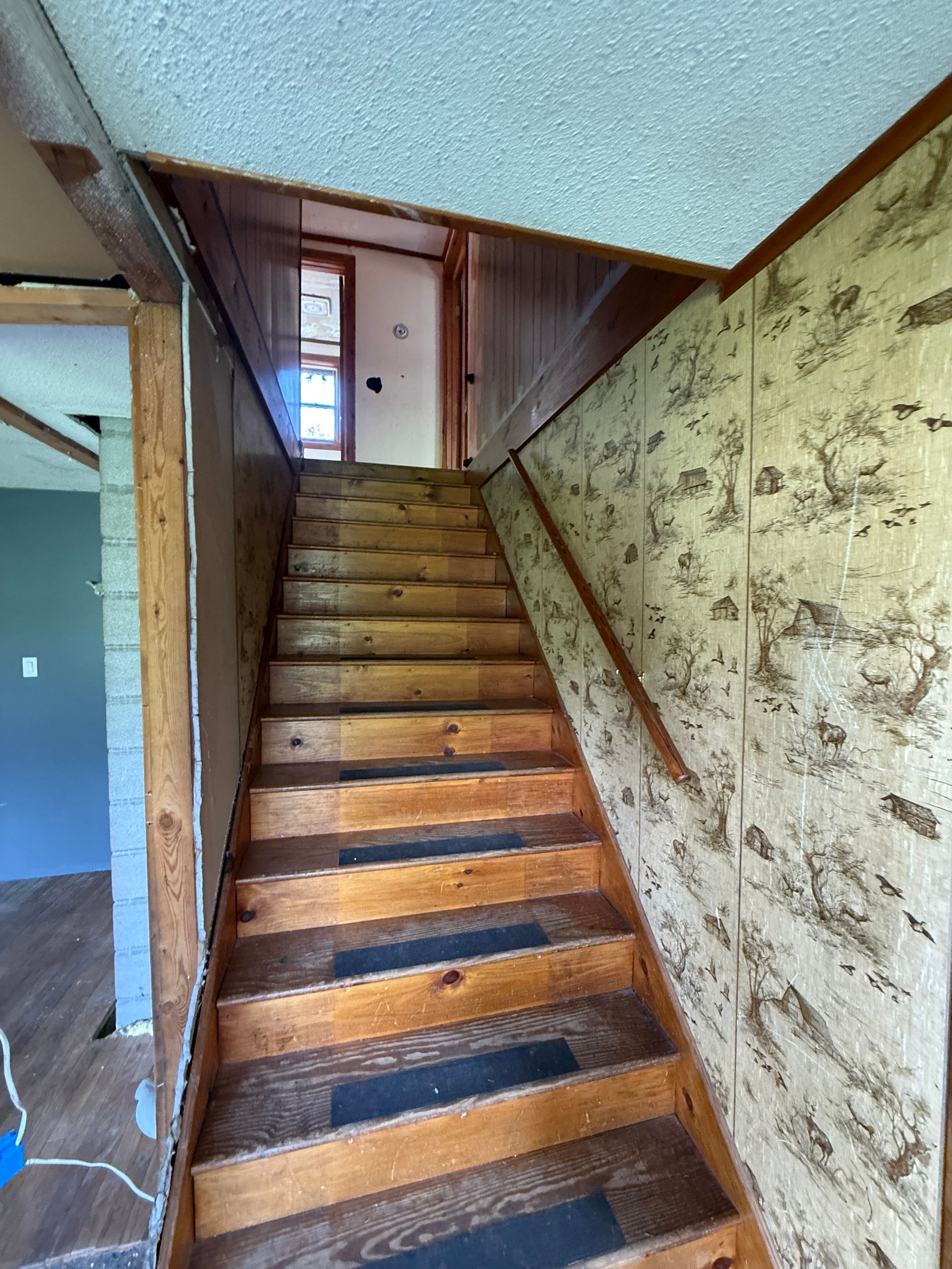 Old wooden staircase in a narrow hallway with patterned wallpaper and a window at the top