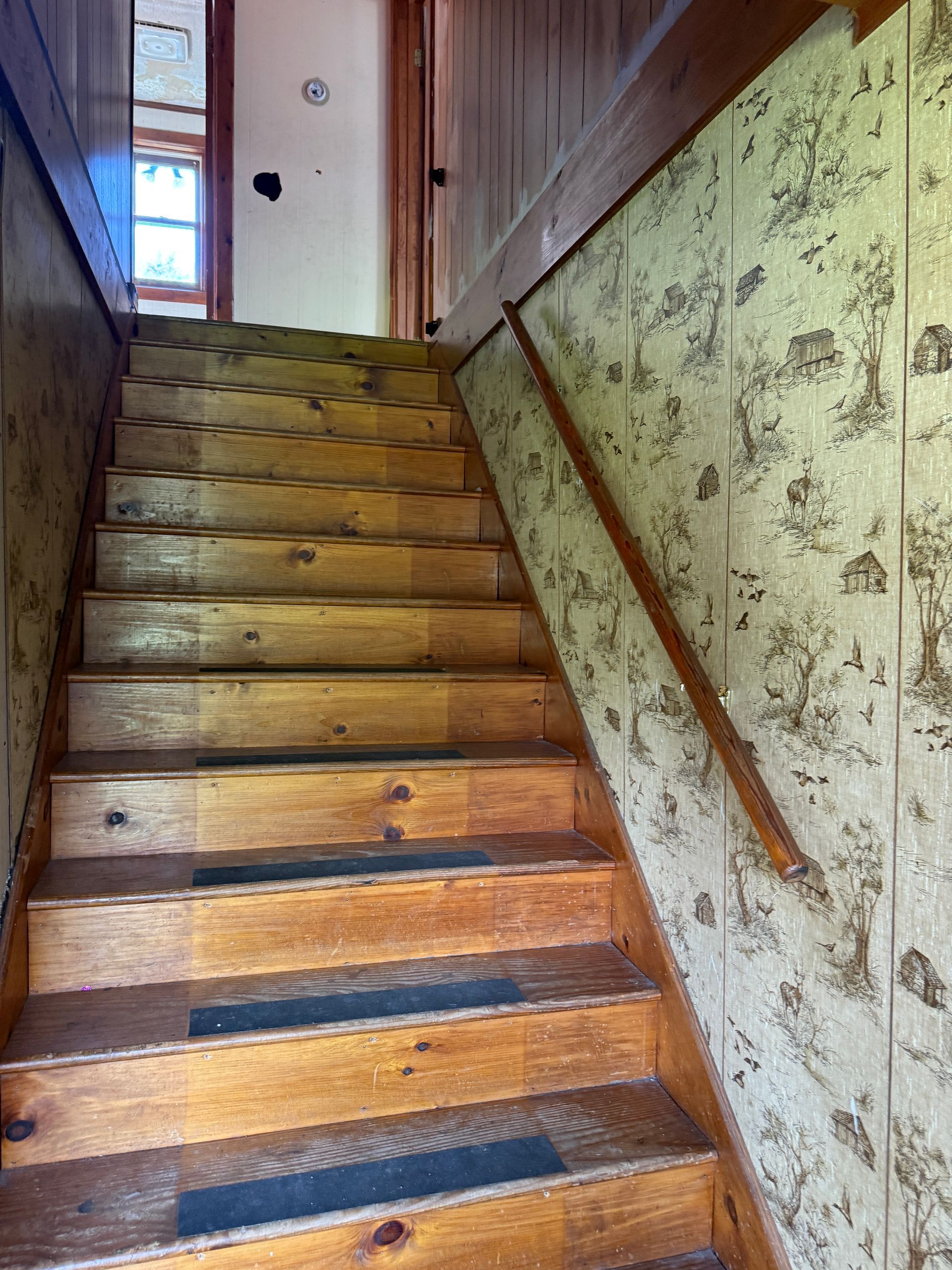 Wooden staircase in a narrow hallway with floral wallpaper and a door at the top.