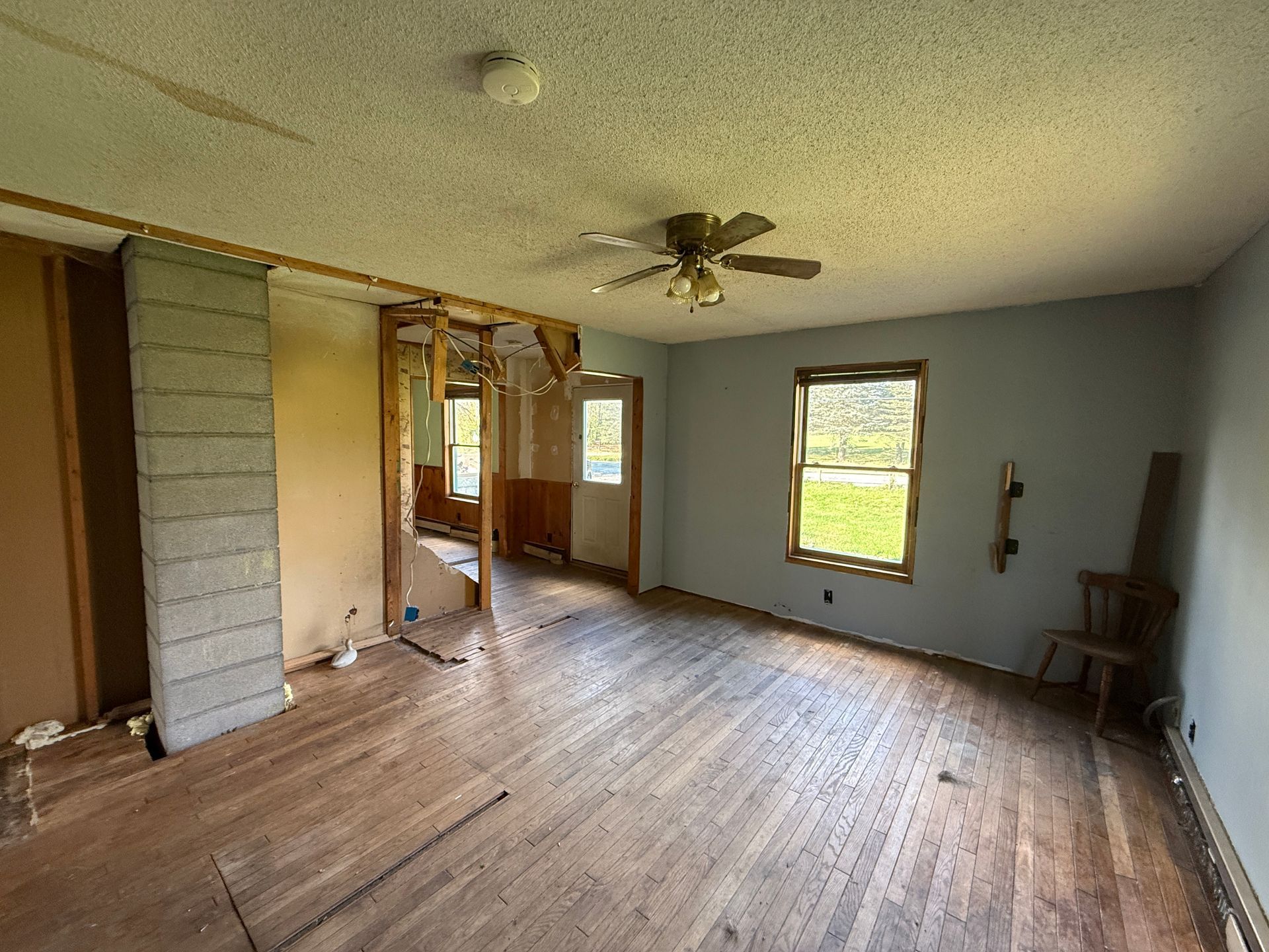 Empty living room with wood floors, gray walls, ceiling fan, and large windows.