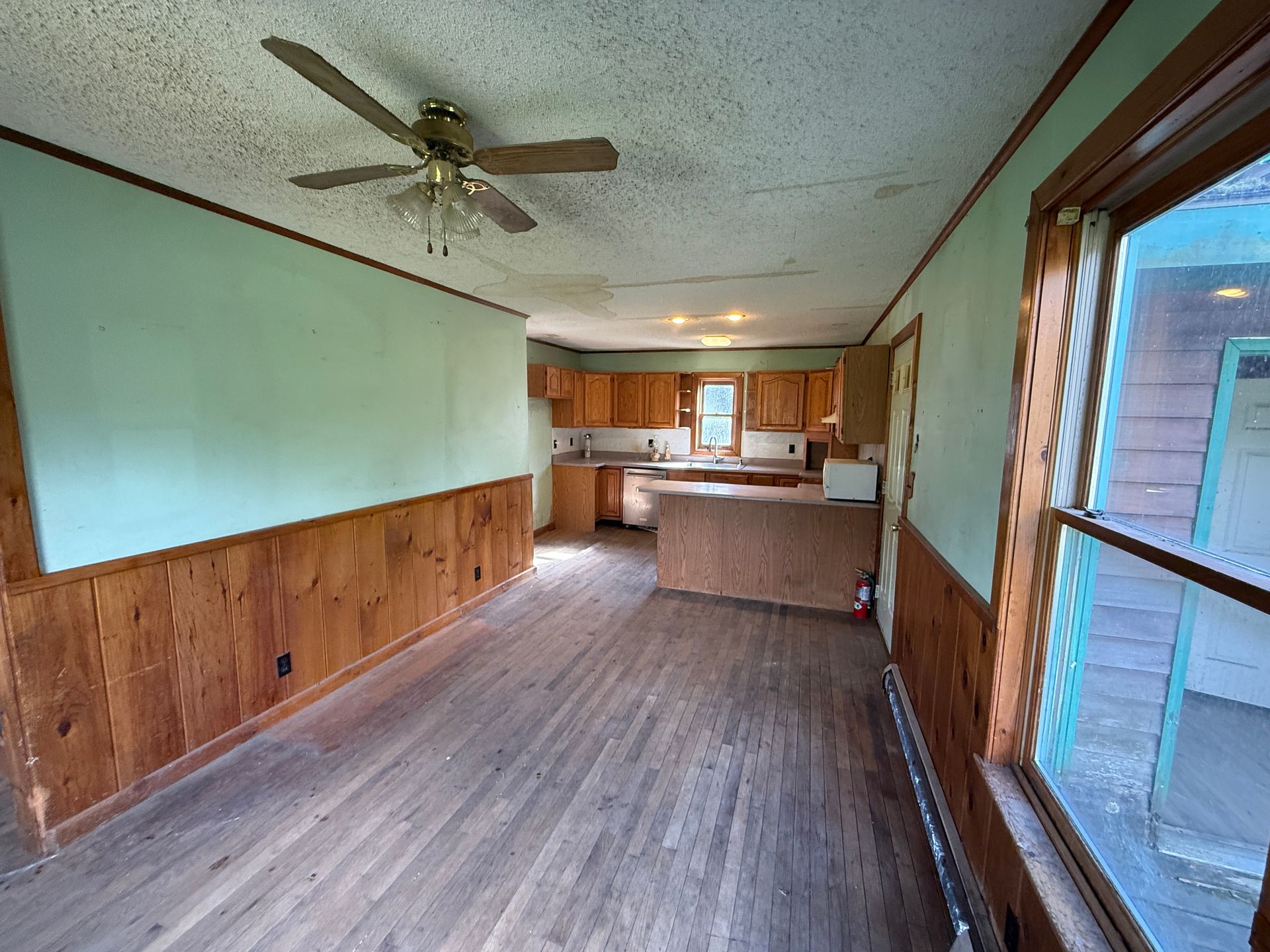 Empty wood-paneled living room with ceiling fan, open kitchen, and sliding glass door.