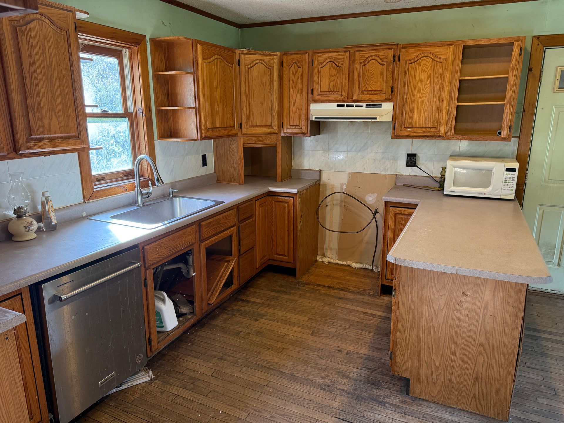 Small kitchen with wood cabinets, stainless dishwasher, and countertop microwave under a window.