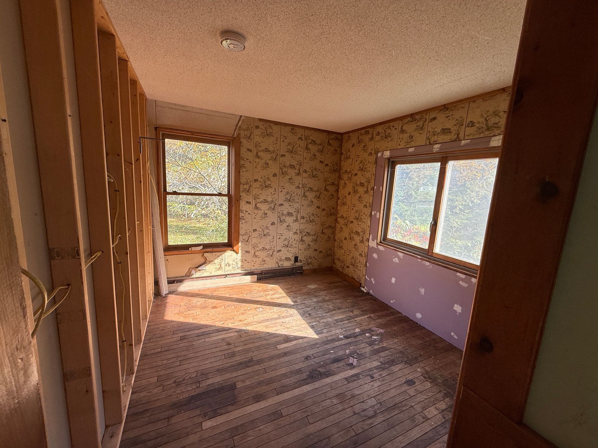 Empty room with worn wood floors, floral wallpaper, and sunlight through large windows.