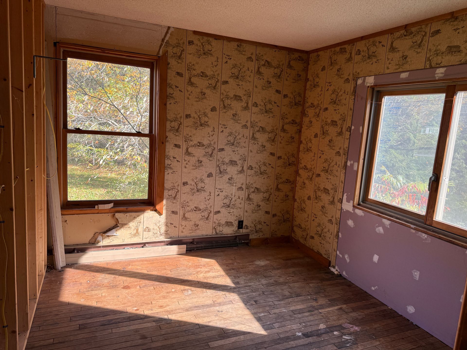 Empty room with floral wallpaper, two windows, and sunlight on bare wooden floor.