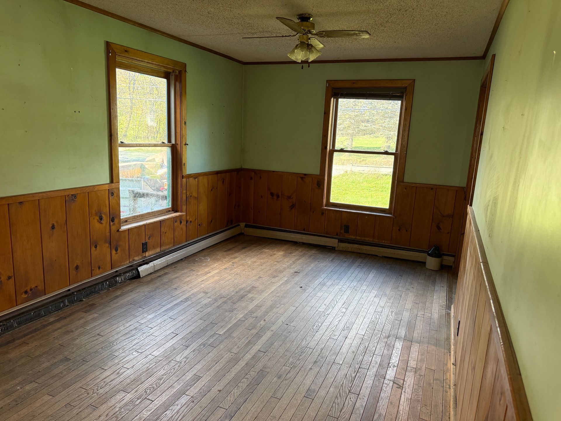 Empty room with wood-paneled walls, green upper walls, and two windows letting in daylight.
