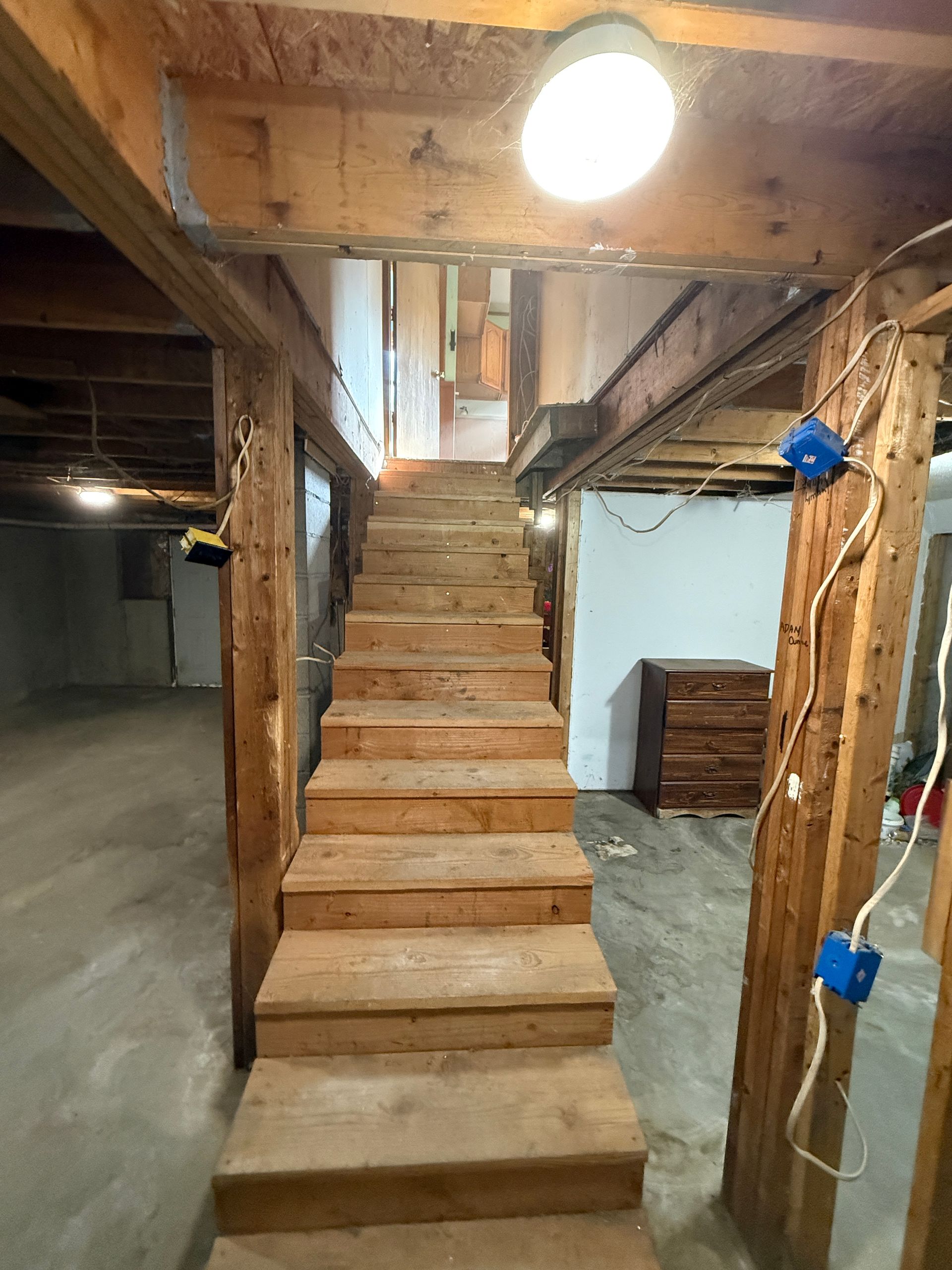 Unfinished basement with wooden staircase leading up under exposed beams and a single bright light.