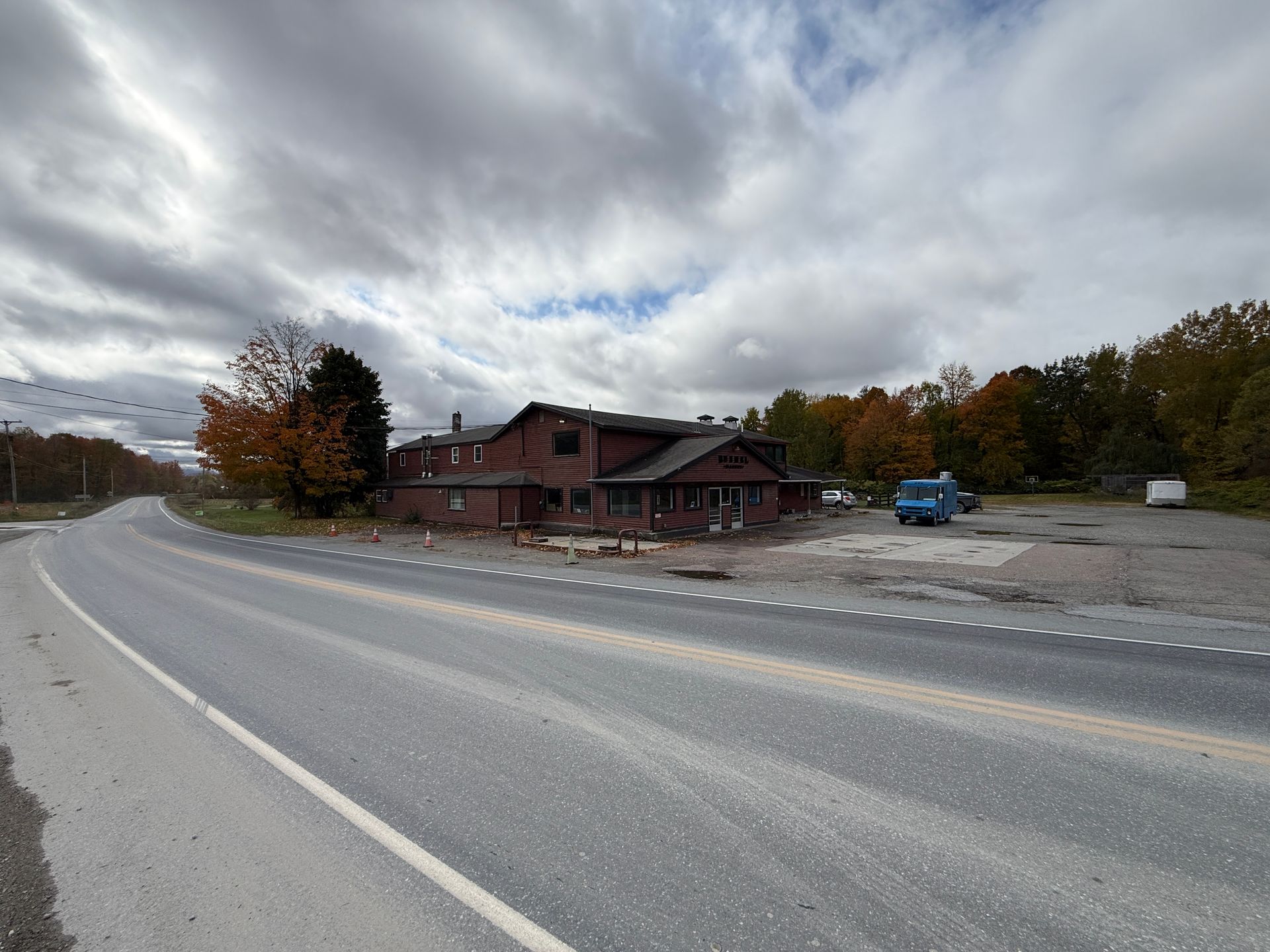 Curving road beside a rustic building and parking area under a cloudy sky, with autumn trees nearby