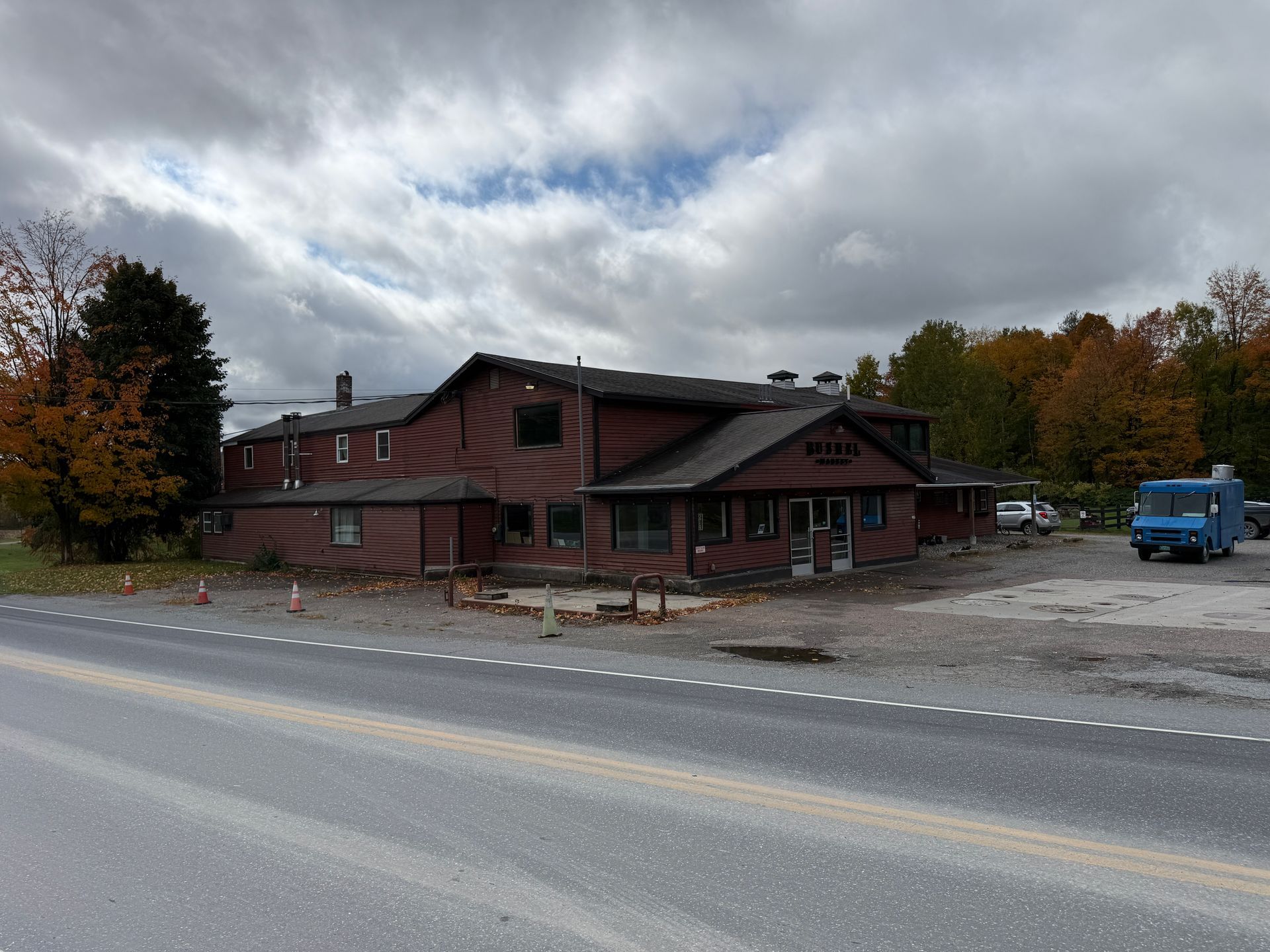Red wooden roadside building under cloudy skies, with a blue truck parked nearby in an autumn setting