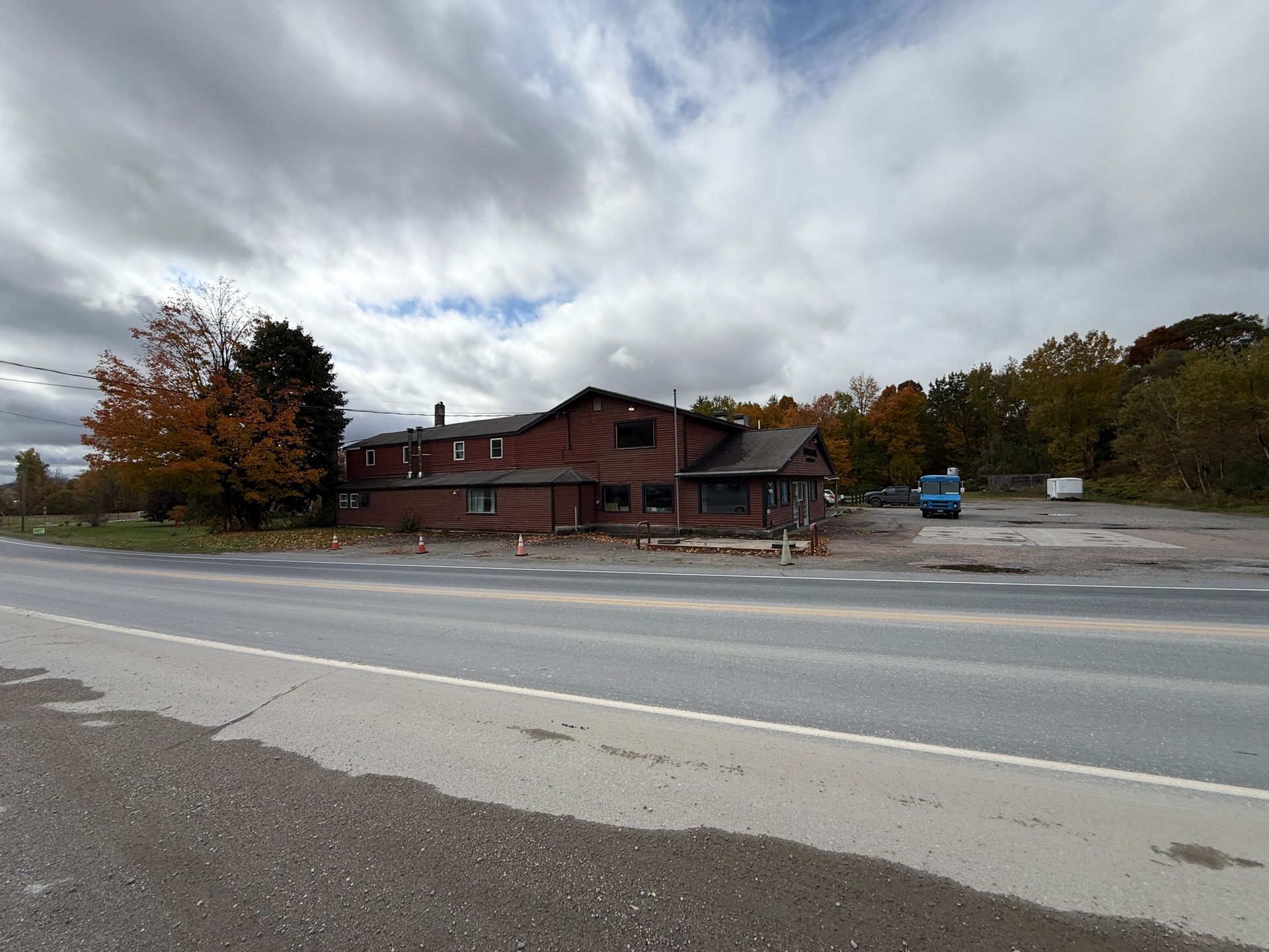 Brick building beside a road under a cloudy sky, with autumn trees and a parking lot nearby.