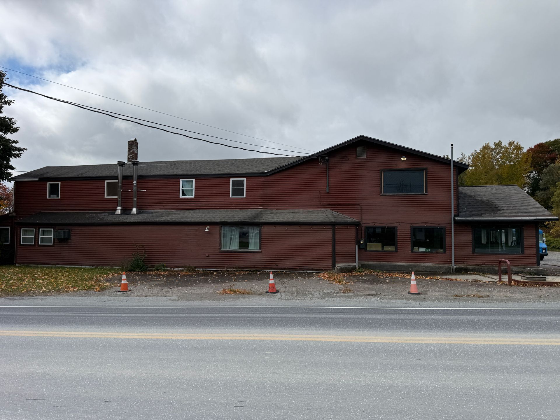Red wooden building with a peaked roof, small windows, and traffic cones along the roadside under a cloudy sky